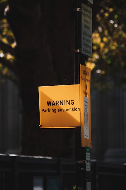 A close-up of a bright yellow warning sign attached to a black metal pole, displaying the text 'WARNING Parking suspension' in black lettering. The sign is positioned at an outdoor street scene during daylight with trees and a fence visible in the background. The sign is illuminated by natural light, highlighting its visibility to drivers and pedestrians. This warning sign is relevant for house removals and moving services provided by Man with Van Stroud Green, as it indicates parking restrictions that can impact the loading and unloading process during home relocation or furniture transport operations.
