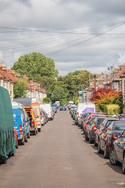 A residential street in Stroud Green with rows of parked cars lining both sides of the narrow asphalt road, including sedans, vans, and small trucks. The scene is captured during daylight with partly cloudy skies overhead. In the background, there are green trees and typical terraced houses with brick facades, some with front gardens and hedges. Overhead, power lines run across the street, and a large green tree dominates the middle distance. The street is part of an area where house removals and moving services, such as those provided by Man with Van Stroud Green, may encounter parking suspensions or restrictions, making careful loading and unloading essential during home relocation or furniture transport operations. The image largely depicts a quiet, suburban setting suitable for residential moves, with no visible people or moving equipment, but the context suggests typical conditions faced during local removals in Stroud Green.