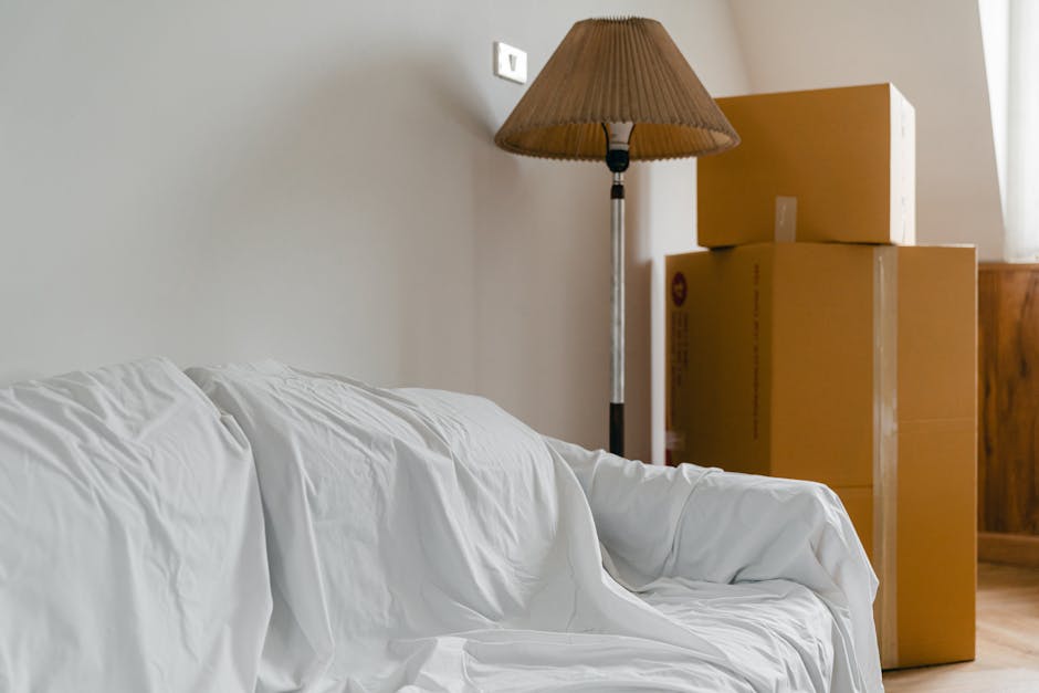 Inside a residential room showing three cardboard boxes stacked near the corner, with two boxes positioned on top of each other and one slightly offset. To the left, a white fabric-covered sofa is partially visible, indicating ongoing packing and home relocation preparations. Behind the boxes, a wooden cabinet is partially seen along with a tall, beige floor lamp with a pleated fabric lampshade. The wall is light-colored, and a small, square electrical socket is visible above the stacked boxes. The scene captures the typical interior environment for furniture transport and packing activities associated with professional removals services offered by Man with Van Stroud Green, supporting house move logistics.
