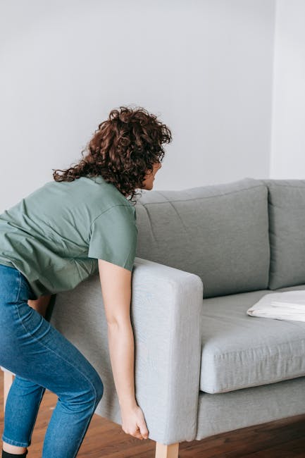 A person with curly brown hair, dressed in a green t-shirt and blue jeans, is in the process of lifting or adjusting a light grey fabric sofa inside a residential living room. The sofa features a single white cushion placed on the seat, and the individual appears to be preparing the furniture for a house removal or transport. The room has wooden flooring and plain white walls, with natural lighting illuminating the scene. This image relates to furniture transport and packing in home relocation, as handled by a professional removals service such as Man with Van Stroud Green, which offers house removal and moving services.
