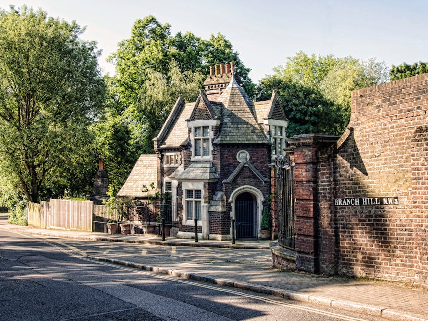A historic, Victorian-style house with a steeply pitched, multi-gabled roof covered in slate tiles, situated on a tree-lined street in Stroud Green. The building features ornate bay windows with white wooden frames, decorative brickwork, and a central arched entrance door painted dark. Surrounding the house is a low brick wall with an iron gate, and a sign on the brick wall reads 'Branch Hill N.W.3'. The street in front has a paved road with double yellow lines and a concrete sidewalk, partially shaded by mature trees with green foliage. The scene captures the quiet residential atmosphere during daylight hours, with natural sunlight illuminating the building's façade and casting shadows. The context of the scene suggests a peaceful setting potentially used for house removals or local relocations, aligning with services such as furniture transport and packing associated with house moves. The company [COMPANY_NAME] might support moving logistics in this area, as evidenced by the neat arrangement of the house and environment prepared for relocation activities.