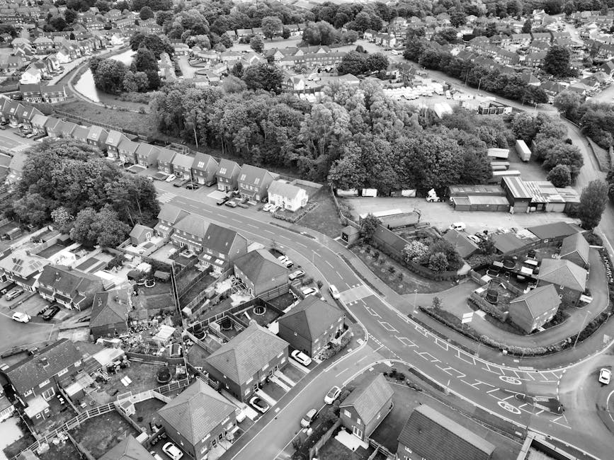 An aerial view of a residential neighbourhood showing rows of semi-detached houses with pitched roofs, arranged along winding streets with parked cars on driveways and roadside. The area includes gree