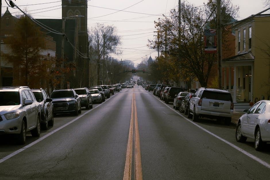 A quiet residential street with parked cars lining both sides, including sedans, SUVs, and station wagons, in various colours such as white, silver, and black. The road has a double yellow line in the centre, indicating no overtaking, and the surface appears slightly worn with minor cracks. On the right side, there are houses with front gardens, some featuring steps leading to their entrances, and a few with visible bay windows. Trees with autumn foliage extend over the pavement, partially obscuring overhead utility wires and street signage, including a vertical banner attached to a pole. The sky is overcast, casting diffuse natural light across the scene. In the context of home relocation and furniture transport, such a street layout is typical for residential removals, with space for loading and unloading. Occasionally, vehicles may be temporarily moved for larger furniture or appliance loading, which is carried out by professional removals services such as Man with Van Stroud Green, especially on streets like this, supporting efficient packing and moving logistics along Ferme Park Road in Stroud Green.