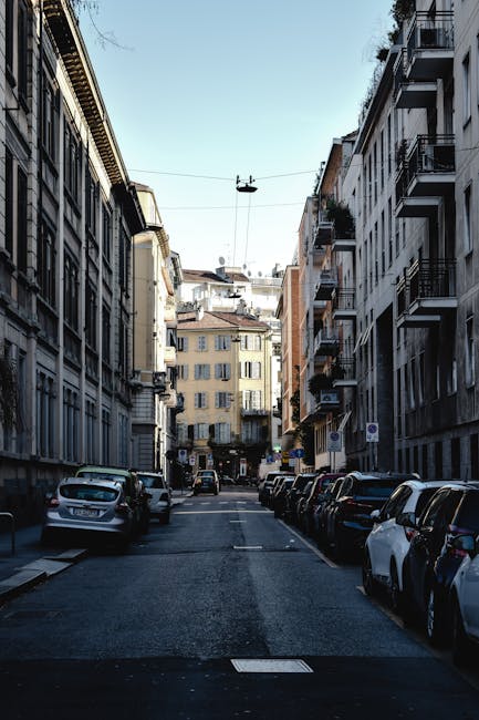 A narrow residential street with multi-storey buildings on both sides, featuring balconies and traditional European-style facades. Parked cars line both sides of the street, including sedans and small vans, with some vehicles partially obscuring the lower sections of the buildings. Overhead, a cable car or gondola system is visible, with cables running across the street and a small cabin suspended in mid-air. The sky is clear, and the scene is illuminated by natural daylight, with shadows cast by the buildings. The road surface appears paved, and the environment suggests an urban setting suitable for house removals and home relocation services. This image could be used to illustrate the challenges of navigating narrow streets during furniture transport or moving logistics, as managed by companies like Man with Van Stroud Green.