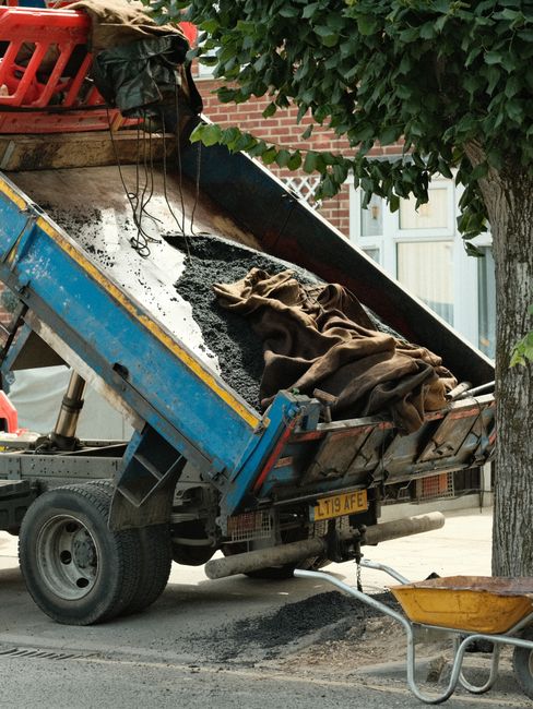 A small blue dump truck used for construction or maintenance is positioned on a residential street with its bed tilted upward, releasing black asphalt onto the pavement. Inside the truck's bed, there are brown fabric tarps and a pile of asphalt material. A yellow wheelbarrow is placed nearby on the ground, ready for material transfer. The background shows a brick building with white-framed windows, alongside a large tree with green foliage. The scene appears to be part of a road maintenance or street improvement project, with the truck part of the moving or construction equipment used in house removals or related services, as provided by Man with Van Stroud Green.