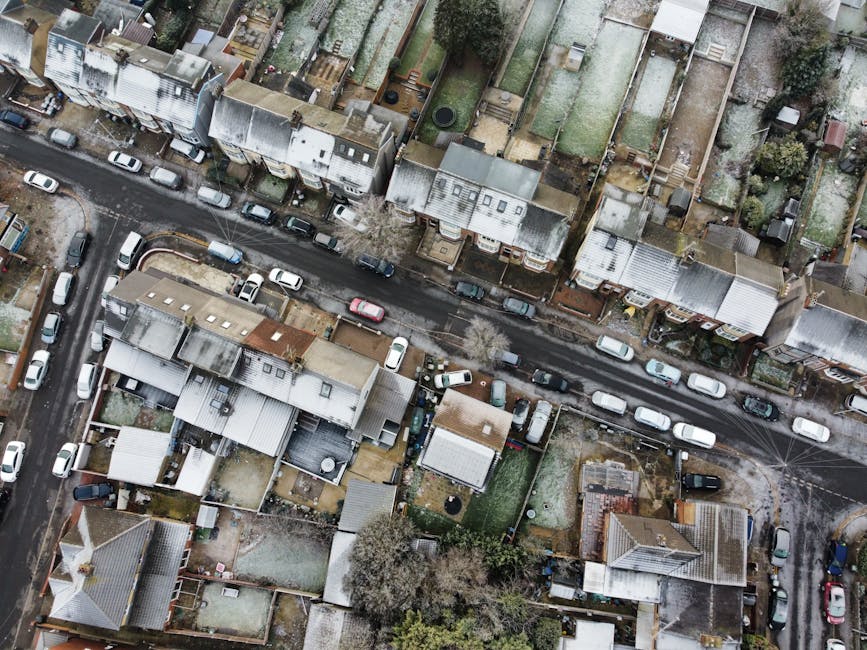 Aerial view of a residential street in Stroud Green showing closely spaced terraced houses with pitched roofs, some covered with metal sheeting. Parked cars are lined along both sides of the street, with a few vehicles occupying the curb space. The street appears to be temporarily used for loading, possibly during a home relocation, with a visible area of activity near the entrance of a property. Several cardboard boxes, plastic wrapping, and furniture are situated on the pavement and in the driveway, indicating packing and moving preparations. A man from Man with Van Stroud Green is engaged in loading furniture and boxes into a van parked adjacent to the house, using equipment such as trolleys and straps to facilitate the process. The surrounding gardens and backyards contain outdoor furniture, garden sheds, and trees, with some areas showing signs of winter dormancy. The overall scene captures the typical logistics involved in furniture transport and packing during house removals in a residential neighbourhood.