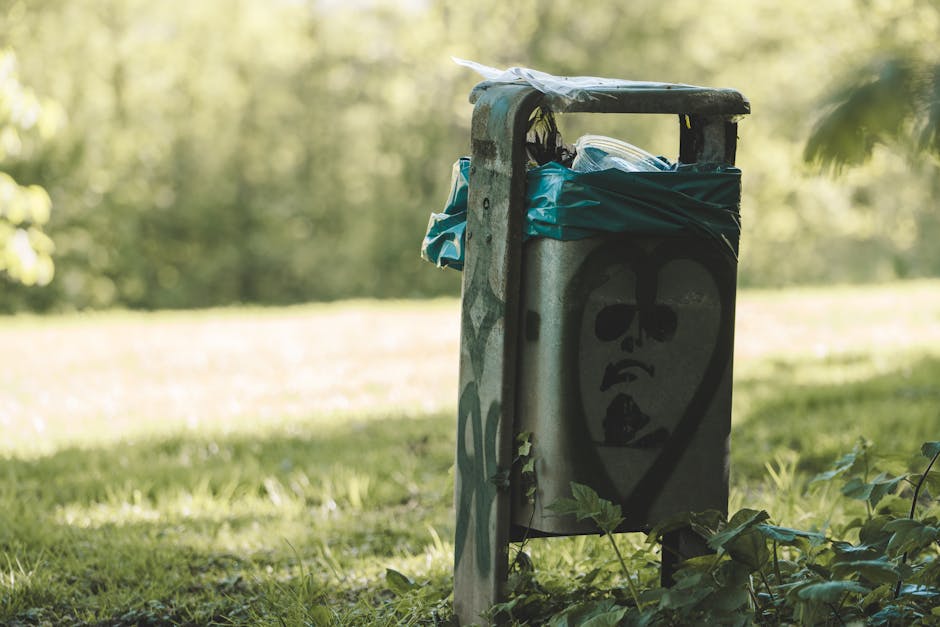 A close-up of a weathered metal waste bin situated outdoors on a grassy area during daylight, with trees and blurred greenery in the background. The bin features a large, black graffiti-style face drawing with sunglasses and a slightly open mouth, and is partially covered by blue plastic wrapping or bags hanging over the top. Surrounding the bin are some small plants and leaves, with the pavement or ground not visible. The scene suggests a typical urban or suburban environment where waste collection or disposal occurs, relevant to house removals and moving services provided by Man with Van Stroud Green, particularly within the context of adhering to Islington Council waste rules for property relocations.