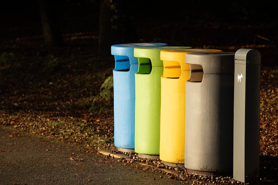 A close-up of a weathered metal waste bin situated outdoors on a grassy area during daylight, with trees and blurred greenery in the background. The bin features a large, black graffiti-style face drawing with sunglasses and a slightly open mouth, and is partially covered by blue plastic wrapping or bags hanging over the top. Surrounding the bin are some small plants and leaves, with the pavement or ground not visible. The scene suggests a typical urban or suburban environment where waste collection or disposal occurs, relevant to house removals and moving services provided by Man with Van Stroud Green, particularly within the context of adhering to Islington Council waste rules for property relocations.