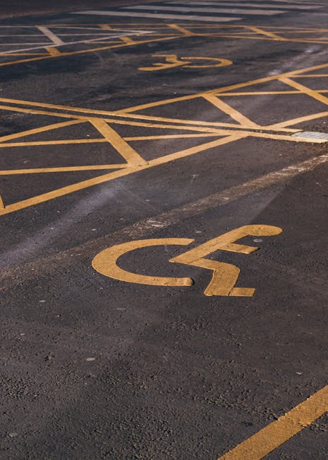 View of a parking lot with multiple yellow wheelchair accessibility symbols and striped markings painted on dark asphalt. The symbols are located near the curb and within designated accessible parking spaces, with some paint slightly worn. In the background, there is a loading area with a curb, and the surface shows signs of recent marking application or cleaning. The scene is well-lit, indicating daytime, and the parking lot appears empty with no vehicles present. This setting reflects an accessible parking area suitable for home relocation and furniture transport when arranging moving logistics, as managed by companies like Man with Van Stroud Green.