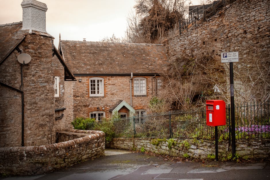 A residential street scene featuring an old stone house with a pitched tiled roof, situated behind a black metal fence and a stone wall. The house has three visible windows with white frames, and a small green-and-white porch roof over the front door. To the right of the house, there is a red post box mounted on a black pole, and a parking restriction sign indicating parking limitations from Monday to Saturday between 8 am and 6 pm, with a note for blue permit holders. The street pavement is visible in the foreground, slightly wet, and the background includes a hillside with some bare trees and a stone retaining wall. The scene suggests a quiet, historic neighbourhood typical of Stroud Green, suitable for residential home relocations or furniture transport activities by [COMPANY_NAME], such as house removals or moving services related to packing and logistics.