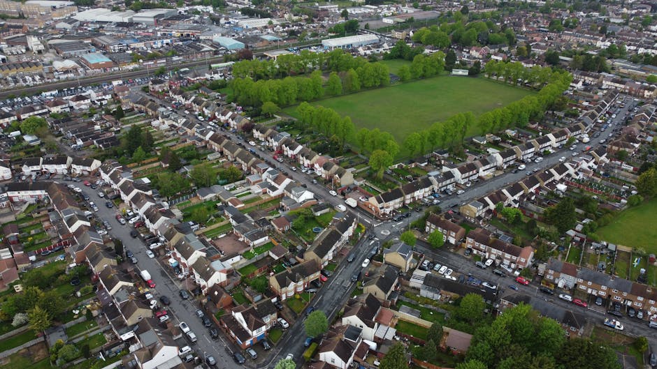 An aerial view of a residential neighbourhood in Stroud Green showing tightly packed terraced houses, some with small back gardens, and narrow streets filled with parked cars. At the center, there is a large green park with evenly spaced trees lining its perimeter, providing open space amid the housing. A bridge crossing over a railway or road runs through the area, connecting different parts of the neighbourhood. The surrounding homes and streets are uniformly arranged, with rooftops varying in color and style. The overall scene captures the dense urban layout specific to a typical UK suburban area, highlighting the proximity of residential properties to communal green space, ideal for home relocation, furniture transport, and packing and moving processes as supported by companies like Man with Van Stroud Green.