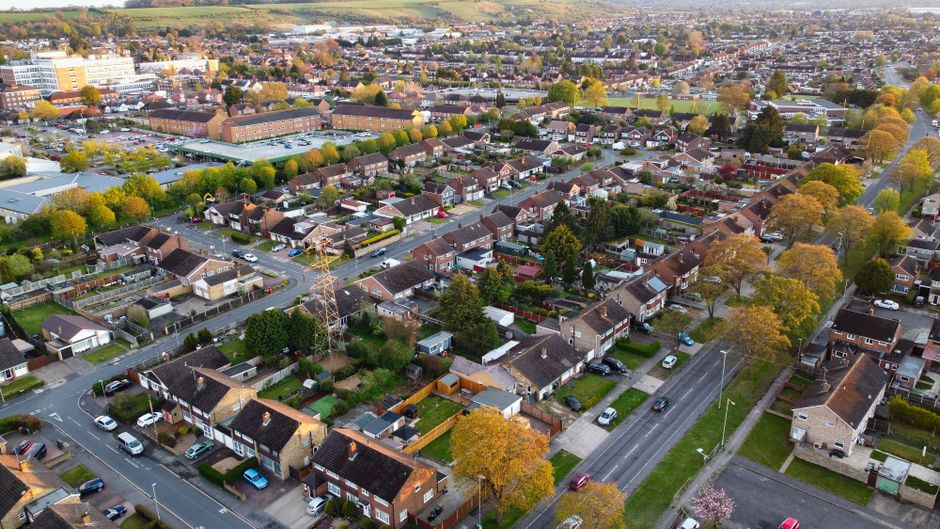 An aerial view of a residential neighbourhood in Stroud Green showing tightly packed terraced houses, some with small back gardens, and narrow streets filled with parked cars. At the center, there is a large green park with evenly spaced trees lining its perimeter, providing open space amid the housing. A bridge crossing over a railway or road runs through the area, connecting different parts of the neighbourhood. The surrounding homes and streets are uniformly arranged, with rooftops varying in color and style. The overall scene captures the dense urban layout specific to a typical UK suburban area, highlighting the proximity of residential properties to communal green space, ideal for home relocation, furniture transport, and packing and moving processes as supported by companies like Man with Van Stroud Green.